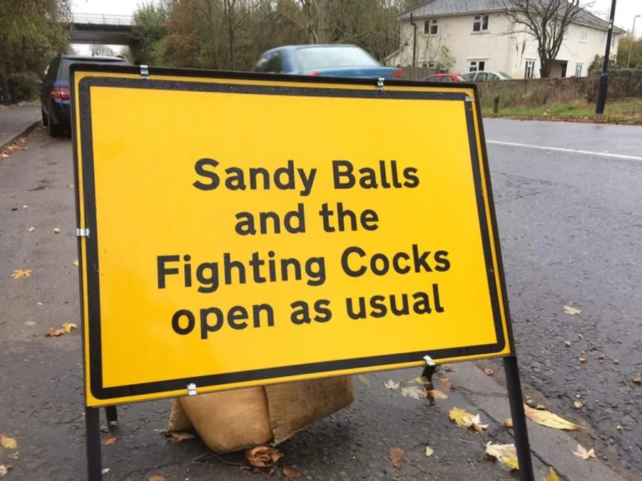Roadside sign stating that Sandy Balls and the Fighting Cocks are still open during road works